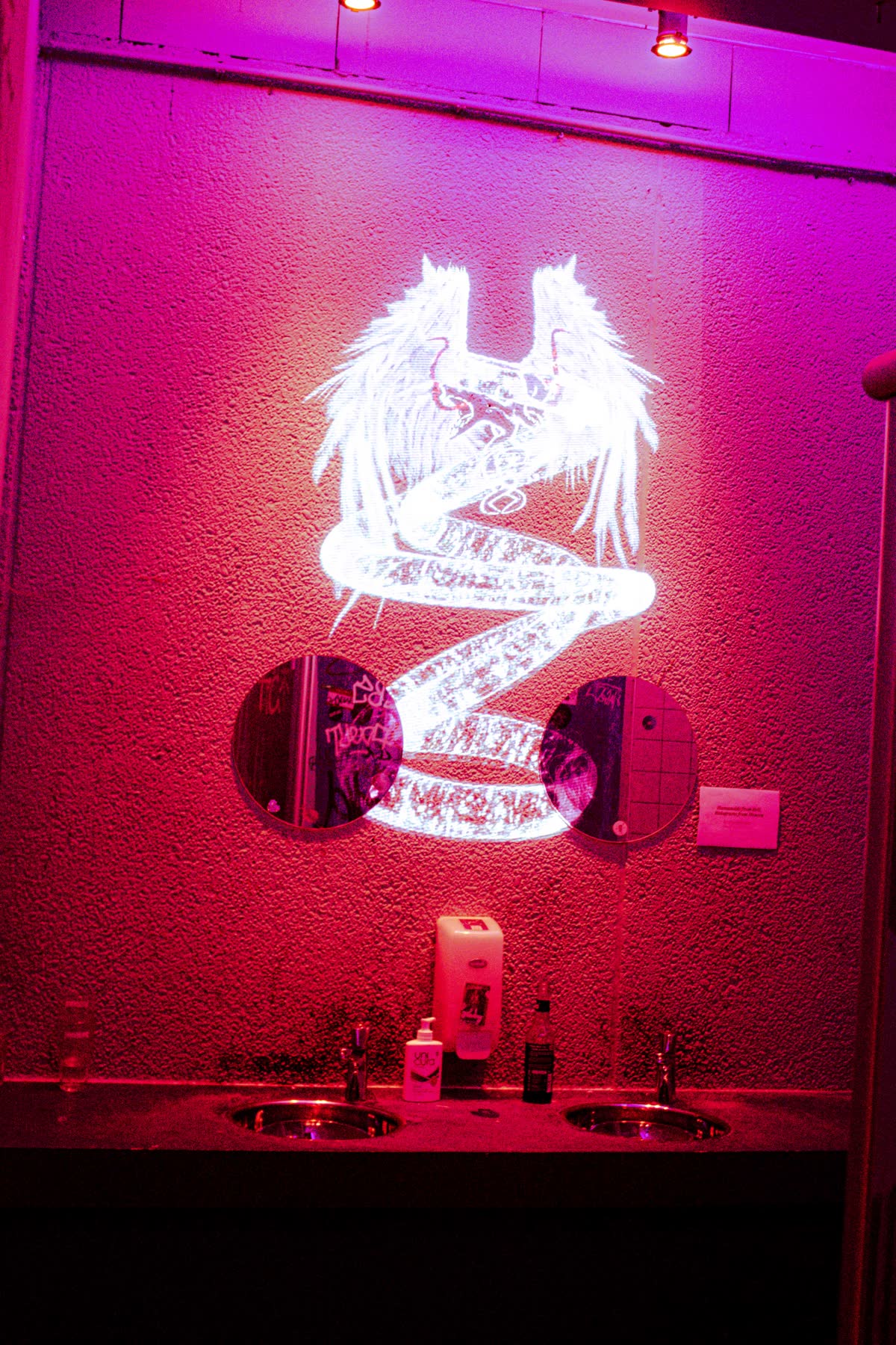 de gevleugelde figuur geprojecteerd op de wc-muur boven de wasbakken, de hele ruimte magenta